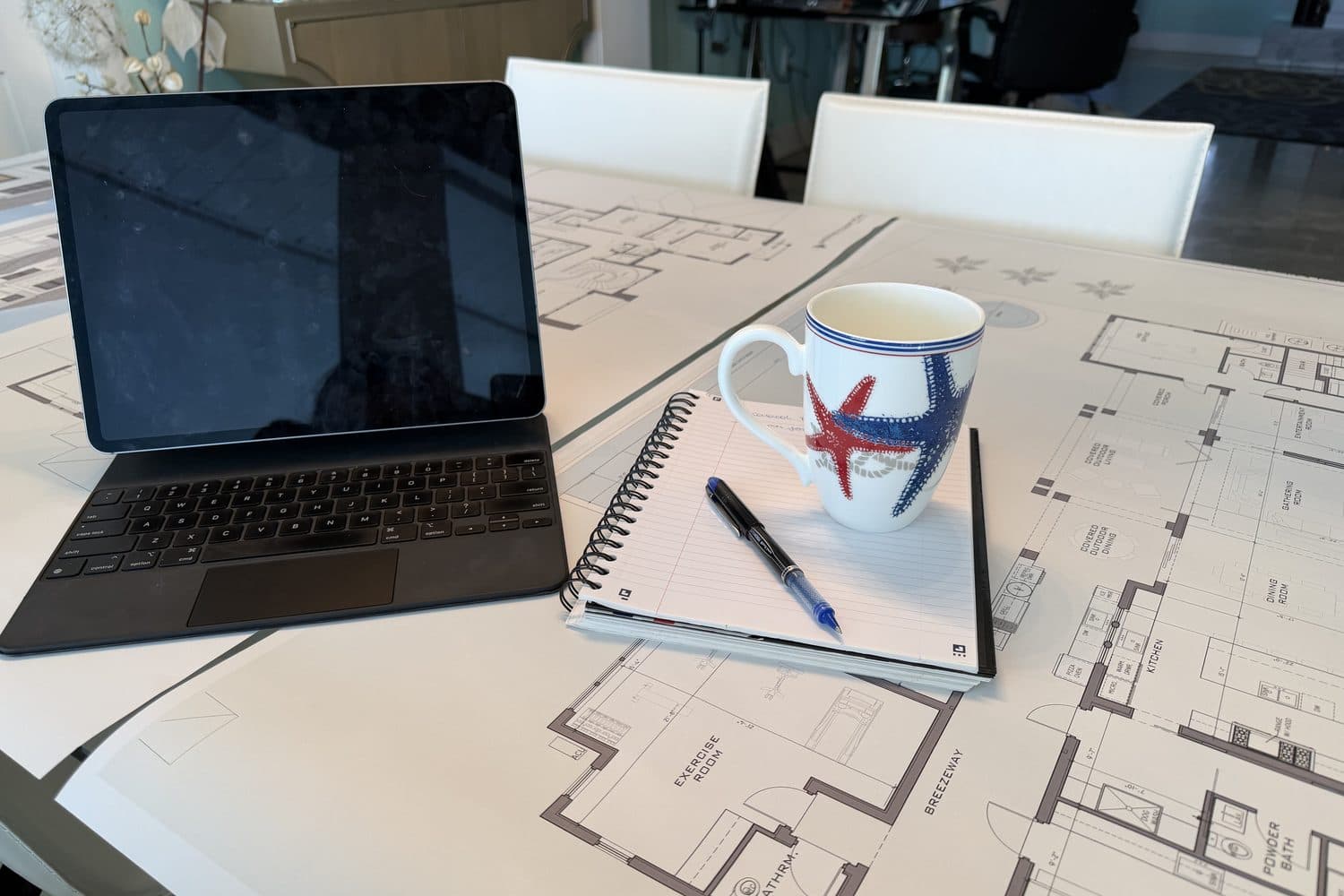 Floor plans spread across a table with a tablet, a notebook, a pen and a coffee mug, ready for a project review.