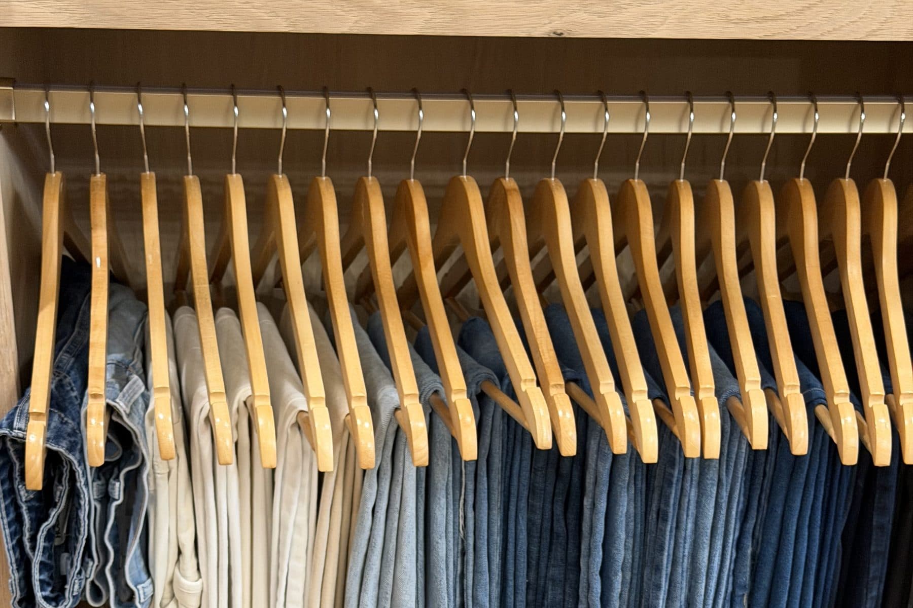 A built in oak closet with a brass rod, matching wooden hangers and jeans hung from light to dark wash.